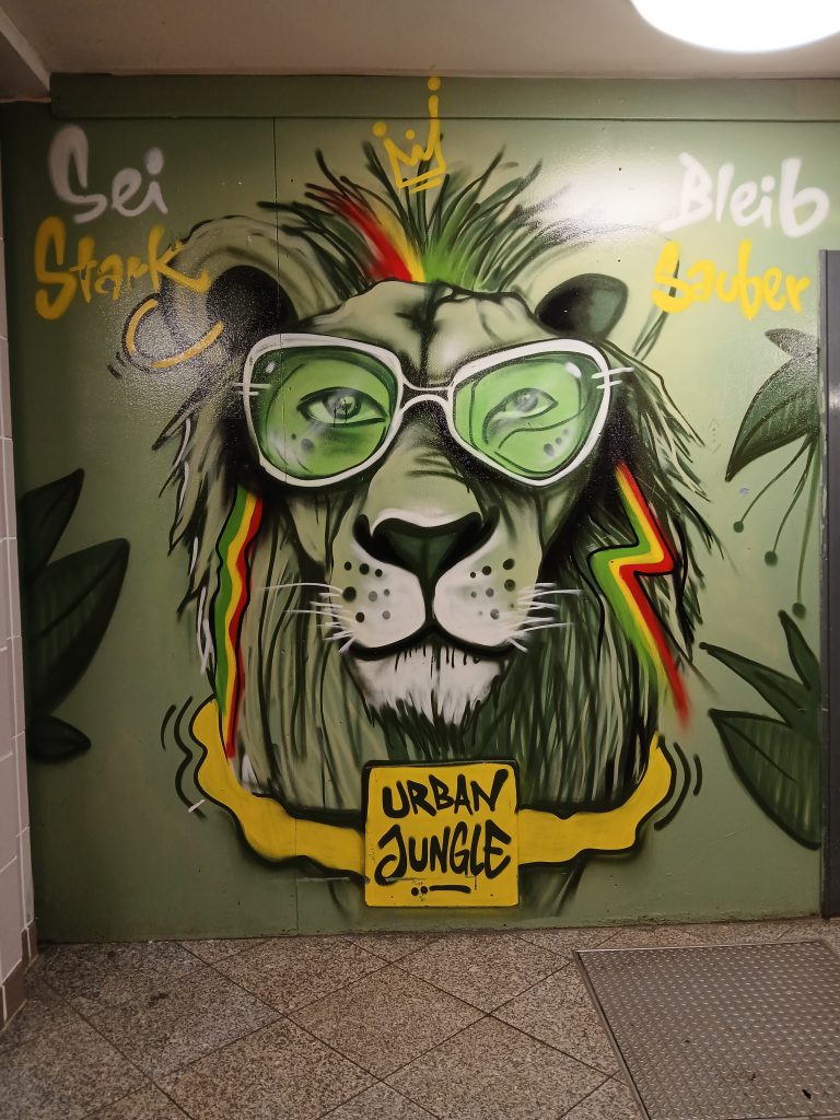 Street art, graffiti, a lion faces you. It has a punk style mane in Jamaican colors green, yellow and red and wears green glasses. It wears an "urban jungle" necklace and caption left reads "Sei stark" (be strong) and "Bleib sauber" (stay clean)